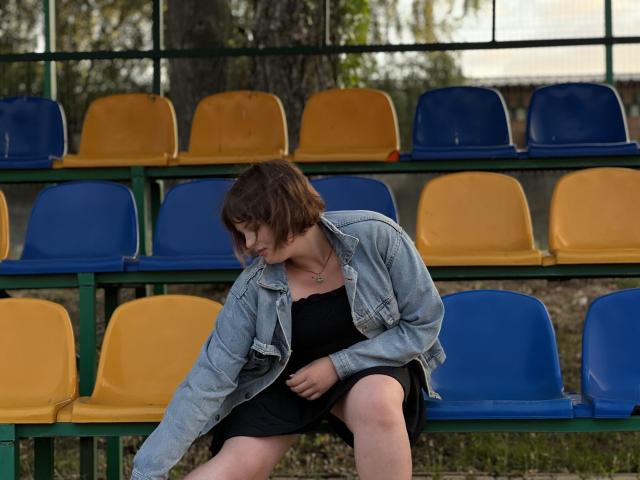 StaceyLex with short brown hair, wearing a denim jacket over a black dress, sits on blue and yellow plastic seats, looking down with a slight frown.