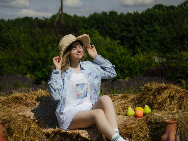 TinaRedly, met blond haar, draagt een strohoed, een blauw shirt, een witte jurk en blauwe sokken, zittend op hooi, met een hoed in haar hand, met groene peren en een oranje appel naast haar..