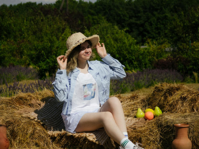 TinaRedly, sitting on hay, wearing a straw hat, blue shirt, white dress, and socks, with her legs crossed, holding her hat, surrounded by apples and a clay pot.