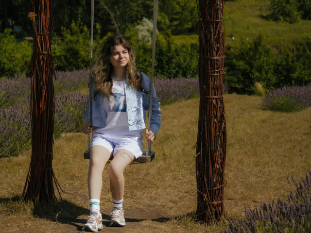 TinaRedly sits on a wooden swing with rusted poles, wearing a denim jacket, white shirt, shorts, and sneakers, with wavy brown hair, in a sunlit lavender field.
