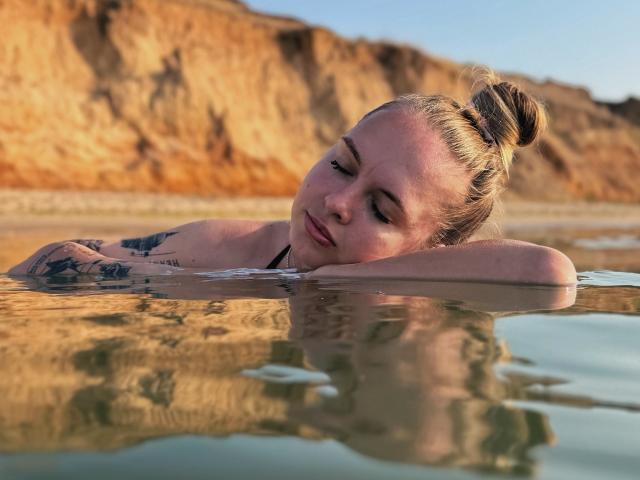 KhaleesiMoon with her head resting on the water, eyes closed, blonde hair in a bun, tattoos on her arms, wearing a black strap, in a sunny, reflective pool.