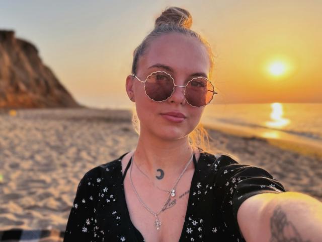 KhaleesiMoon with a bun, wearing round sunglasses, black star-patterned shirt, and necklace, has a crescent moon tattoo below her collarbone, standing on a sandy beach at sunset.