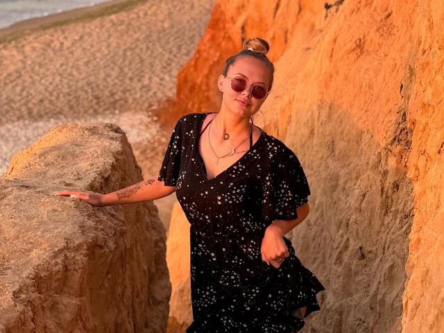 KhaleesiMoon with tan skin, tied-up bun, pink sunglasses, black speckled dress, tattoos on right arm, gold necklace, standing against orange rock, touching gray rock.