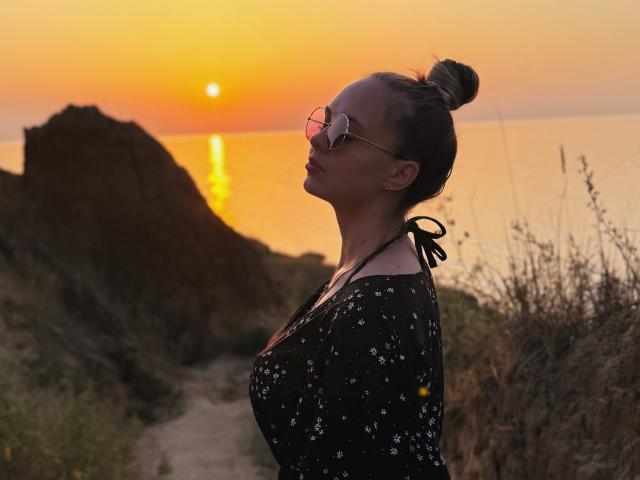KhaleesiMoon with tits in a black floral top, hair in a bun, sunglasses, standing on a path at sunset, looking up, orange sky, sun reflection on water.