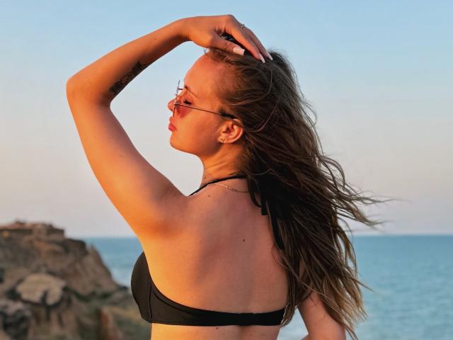 KhaleesiMoon with long brown hair, wearing a black bikini top, raising her right arm, showing her back and small ass, standing by the blue ocean at sunset.