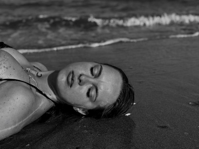 KhaleesiMoon lies on the beach, wet hair, eyes closed, bikini top, sand on her skin, necklace, gentle waves in the background, black and white, relaxed, sensual.
