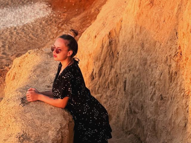 KhaleesiMoon leans against a rock, wearing a black dress with white dots, sunglasses, and her hair in a bun, bathed in warm orange sunlight. Her ass is slightly visible.