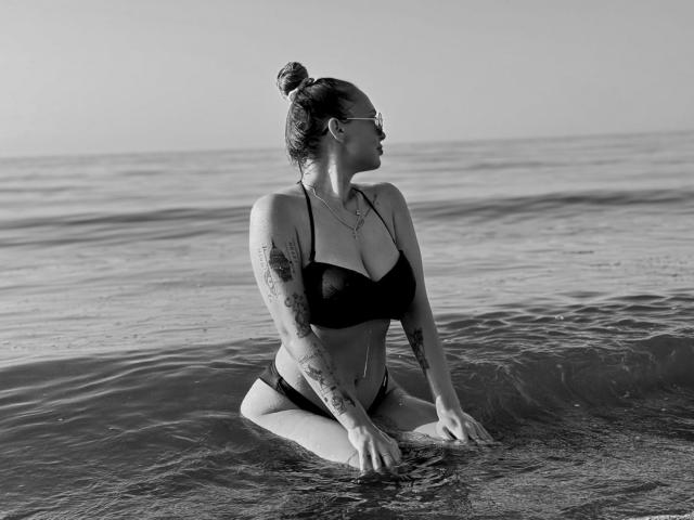 KhaleesiMoon is kneeling in the water, wearing a black bikini, with tattoos on her arms and a bun, looking to the side, in a black-and-white photo.