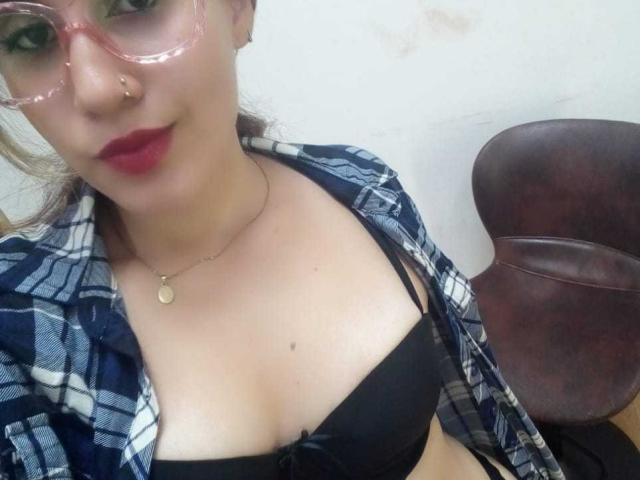 Keidy69 with pink glasses, red lips, nose ring, plaid shirt, black bra showing her tits, and a necklace, sitting in a brown leather chair.