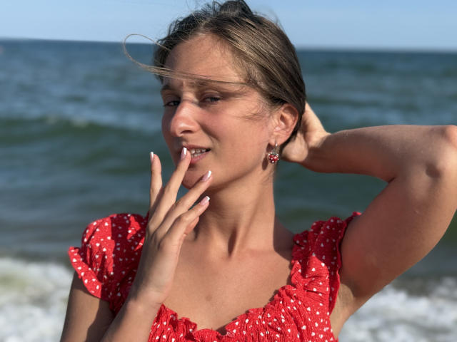 MichelleHotX69 has tanned skin, brown hair, and wears a red polka-dot dress with puffed sleeves. She touches her lips with her fingers, looking sexy with a hand in hair.