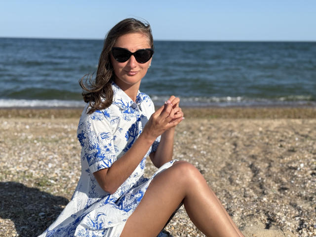 MichelleHotX69 with brown hair, wearing black sunglasses, blue and white floral dress, sitting on pebble beach, hands together, legs crossed, smiling, sunny day, ocean in background.
