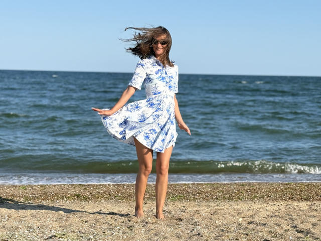 MichelleHotX69, barefoot, wearing a white blue floral dress, standing on a pebbly beach, dress blowing, brown hair, sunglasses, smiling, ocean in background, clear blue sky.