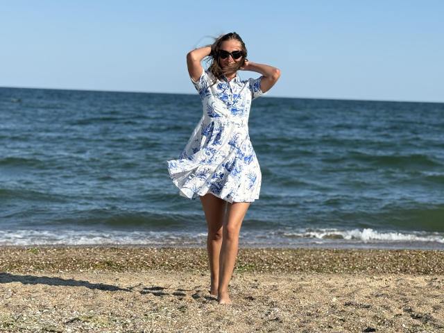 MichelleHotX69 standing barefoot on a pebbly beach, wearing a blue floral dress, sunglasses, and hands in her hair, with a calm ocean in the background.