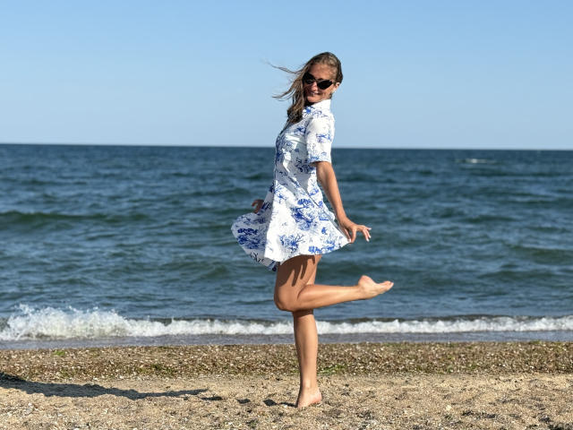 MichelleHotX69, barefoot, standing on sandy beach, white floral dress, blue eyes, brown hair, sunglasses, one leg raised, blowing in wind, ocean, clear sky, smiling, sunny day.