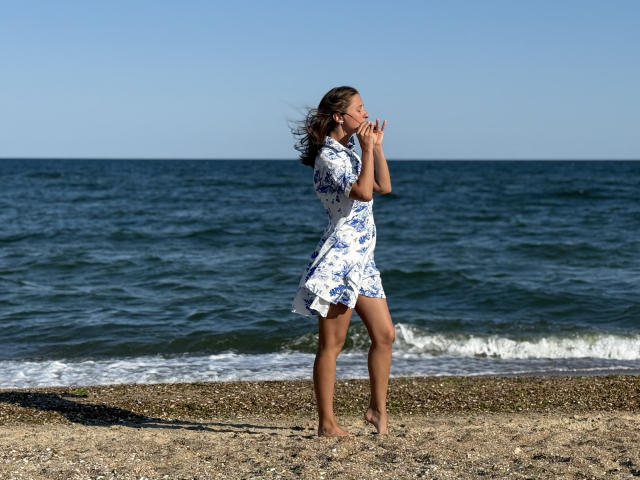 MichelleHotX69 in a blue and white floral dress, standing barefoot on a pebbly beach, wind blowing through her brown hair, arms raised, ocean in the background.