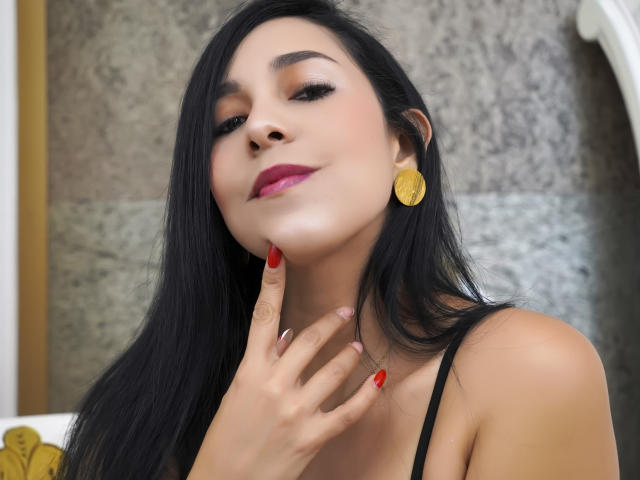 Katty Rivera, con cabello negro largo, lápiz labial rojo y aretes dorados, tocándose la barbilla con uñas pintadas de rojo, mirando hacia arriba con una ligera sonrisa. Se ven los senos, lleva un top negro con tirantes..