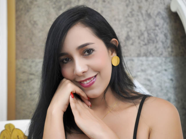 KattyRivera with long black hair, smiling, pink lipstick, yellow earrings, black strap dress, red nail polish, hand on cheek, soft light, pretty face, cute, sexy, relaxed.