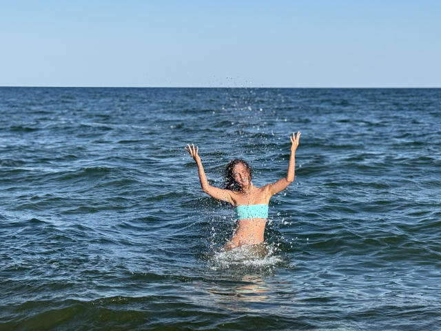 MichelleHotX69's blue bikini top, wet, with arms raised, splashing water around her, standing in a greenish-blue ocean, sunlight reflecting on her skin.