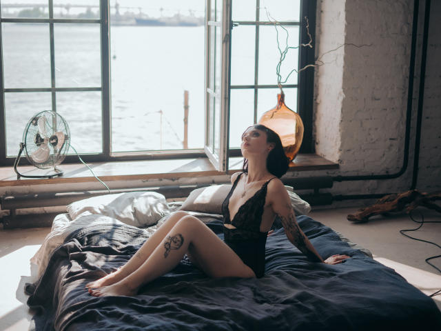HollyVex está sentada em uma cama, vestindo lingerie de renda preta, com os olhos fechados e a cabeça inclinada para trás, exibindo tatuagens na coxa e no braço, com os seios levemente visíveis..