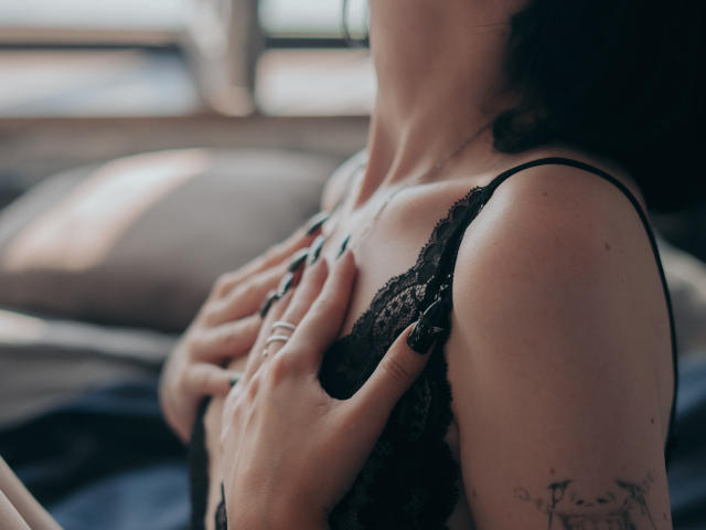 Les mains de HollyVex agrippent ses seins, recouverts d'un soutien-gorge en dentelle noire, ses doigts ornés d'une bague effleurent sa poitrine, révélant des tatouages sur son bras, dans une lumière douce et naturelle..