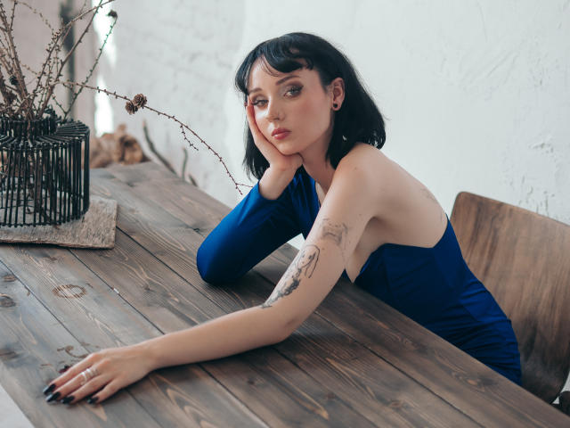 HollyVex, con cabello negro, vestido azul y tatuajes, se apoya en una mesa de madera, con la mano en la barbilla, luciendo seria, con esmalte de uñas negro, una silla de madera, un jarrón de alambre negro y una pared blanca..