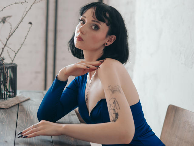 HollyVex avec une coupe de cheveux courte noire, une robe bleue, les seins apparents, un tatouage sur le bras droit, une main sur le cou, une table en bois, un vase noir, une lumière douce, une peau pâle, sexy, un maquillage subtil, un regard confiant..