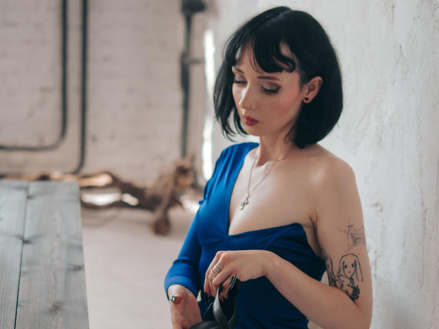 HollyVex con cabello negro corto, vestido azul, hombro descubierto, tatuaje en el brazo superior, sosteniendo gafas negras, mirando hacia abajo, pared de ladrillos blancos, luz suave, casual, sexy, sutil..