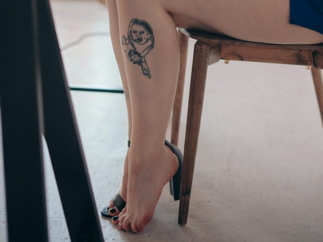 Het been van HollyVex met een zwarte en grijze tatoeage van een verdrietig meisje dat een pop vasthoudt, zittend op een houten stoel, met zwarte hakken en geverfde teennagels..