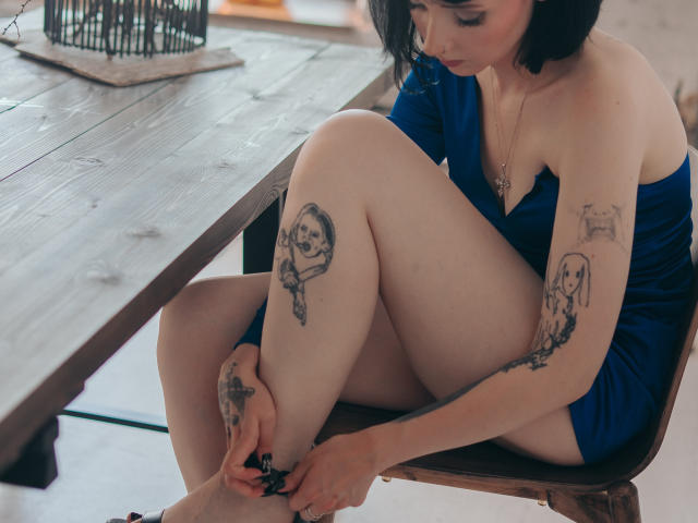 HollyVex with black bob, blue off-shoulder dress, tattoos on arms and legs, sitting on wooden chair, adjusting black ankle bracelet, legs crossed, wooden table in background.