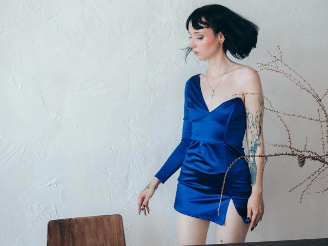 HollyVex with black hair, wearing a blue off-shoulder mini dress, showing a tattoo on her right arm, standing next to a wooden chair, looking to the side.