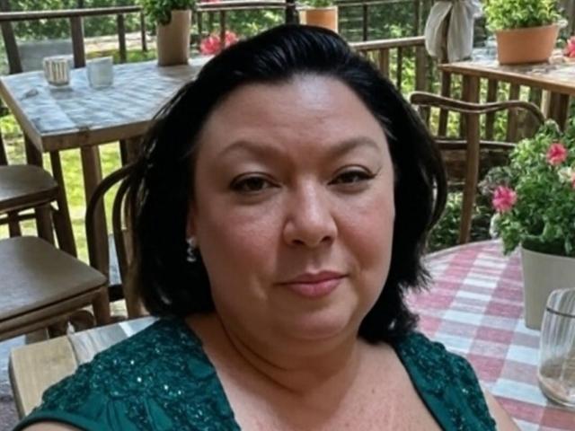 ZarraX, a chubby woman with black hair and green eyes, wearing a green lace top, has small tits and is smiling slightly. She's sitting at an outdoor wooden table.