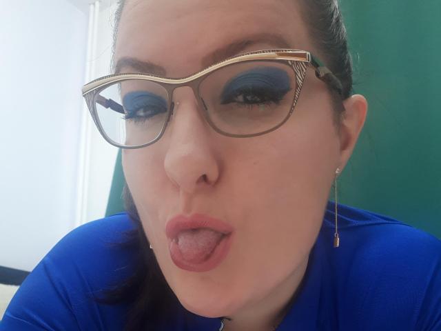 BadAngel with blue eyeshadow, gold-framed glasses, and tongue out, wearing a blue shirt, has light skin, dark hair, and small earrings, looking slightly up.