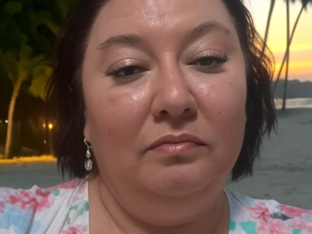 ZarraX has a shiny, slightly sweaty face with subtle makeup, dark short hair, and a floral shirt, wearing a silver earring, looking slightly bored.