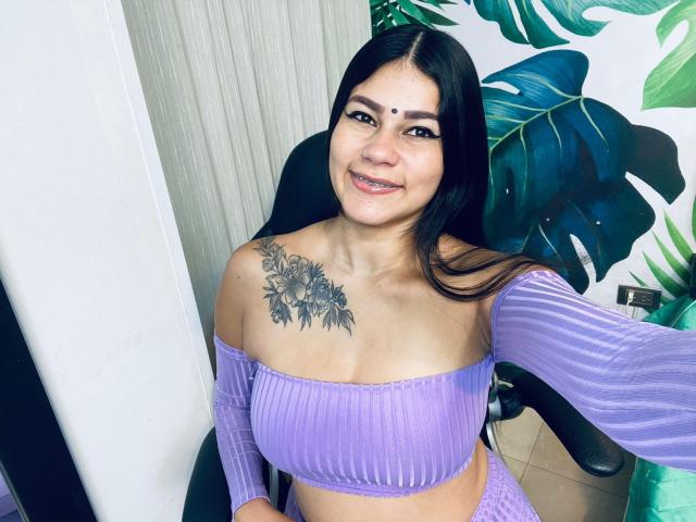 KeraniMarala with long black hair, purple off-shoulder top, and floral tattoo on her right breast, smiling at the camera. Her tits are medium, and she has a cute face.
