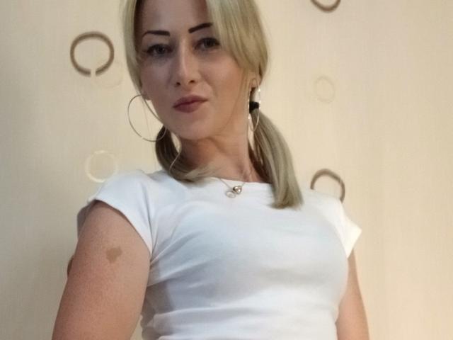 LeilahotX with blonde pigtails, white tee, hoop earrings, subtle makeup, and a small bruise on her right arm, looking slightly bored.