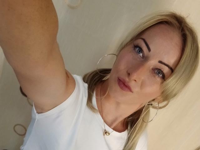 LeilahotX with blonde hair, blue eyes, and hoop earrings, wearing a white shirt, showing a slight pout, taking a selfie, arm raised, with a neutral expression.