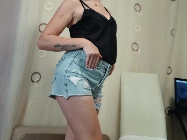 LeilahotX in a black lace camisole and ripped denim shorts, standing side-on, showing a tattoo on her right forearm, with a hand in her pocket.
