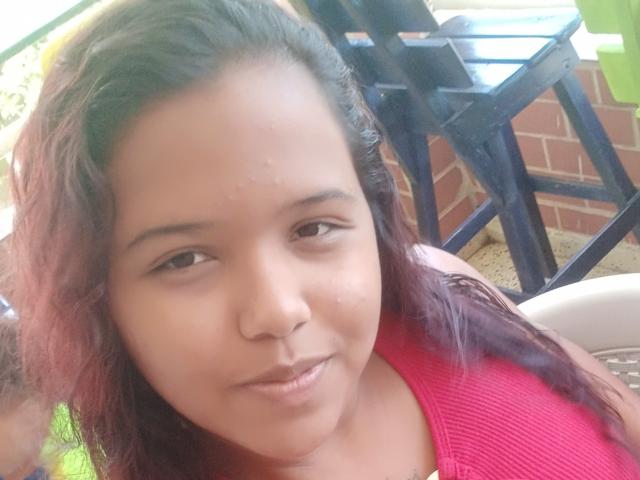 Katy69X has medium brown skin, small nose, dark brown eyes, and wavy purple-red hair. She wears a red top, looking slightly pouty with a small smile.