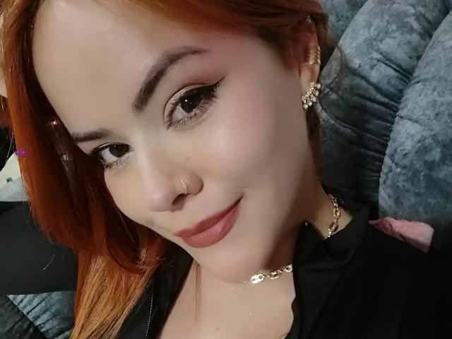 EmilyWattsson has light skin, red hair, and is smiling with eyeliner and mascara. She's wearing gold earrings and a chain necklace, with a black shirt. Boobs are partially visible.