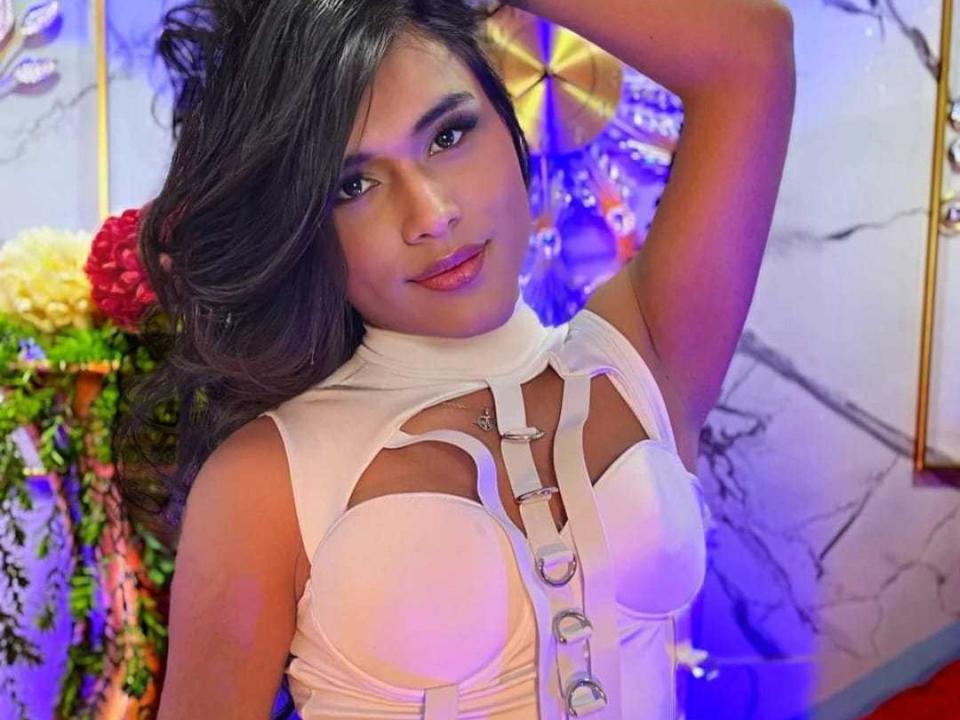 NatashaDown with long dark hair, wearing a white top with straps over her boobs, has a seductive smile, raising her arm. Her makeup is smoky, eyes are half-open.