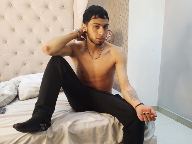 Phoenii69X, shirtless with a toned, tan, and tattooed upper body, black pants, and socks, sits on a bed with one leg up, hand on chin, looking at camera.