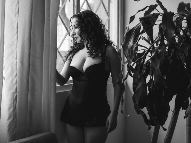 SamanthaAntolin stands in black lingerie, her curly hair cascading, gently touching a curtain, with a potted plant to her right, in a black-and-white shot.