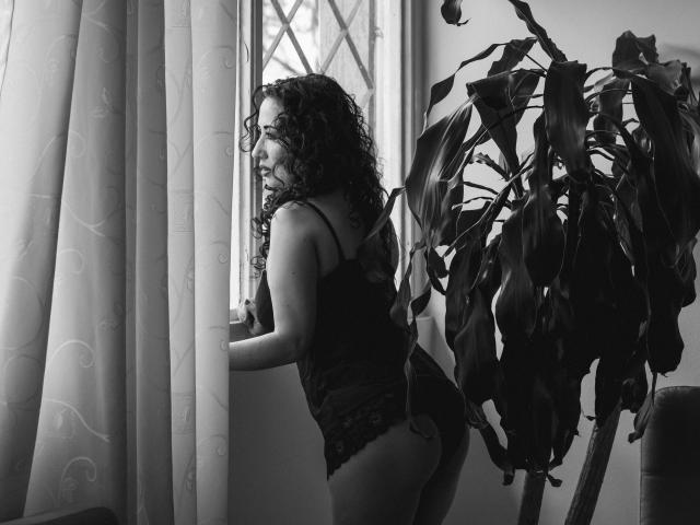 SamanthaAntolin with curly hair, wearing a black lace lingerie set, stands by a window, her ass slightly raised, looking outside, with a large plant on the right. Black-and-white photo.