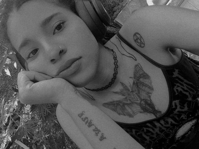Black-and-white selfie of KoralineMiller with headphones, a pentagram tattoo on her shoulder, a bat tattoo on her chest, and "FATPAT" tattoo on her forearm. She's pouting slightly.