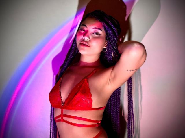 SammyHills69 in red lace bralette, purple braids, nose ring, and hoop earrings, arms raised, showing small tattoos, with pink and purple light casting shadows on her.