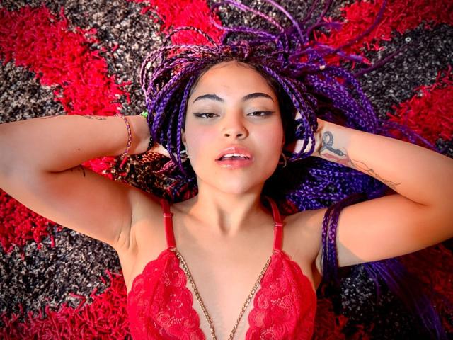 SammyHills69 with purple braids, red lace top, arms raised, tattoo on right forearm, lying on red and black textured surface, looking at camera with slightly parted lips.