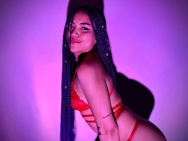 SammyHills69 with long blue braids, red lace bra and strap, smiling, showing off her ass and small tattoo on her arm. Purple lighting. She's cute with a sexy smile.