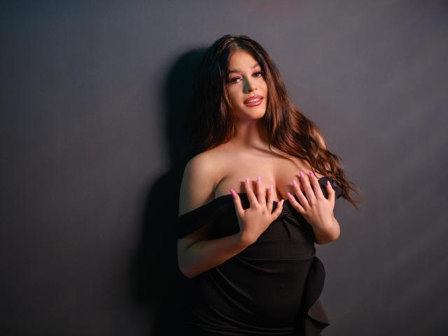 BellaNoirre, avec de longs cheveux bruns, souriante, portant un haut noir à une épaule, les mains couvrant ses seins, ongles roses, debout devant un mur sombre..