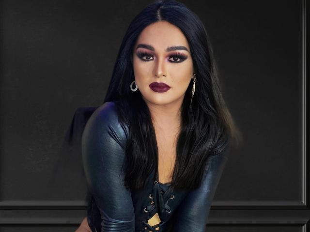 GoddessAnitaX with long black hair, dark purple lipstick, and a tight black leather top, looking seductively into the camera, wearing dangling earrings.