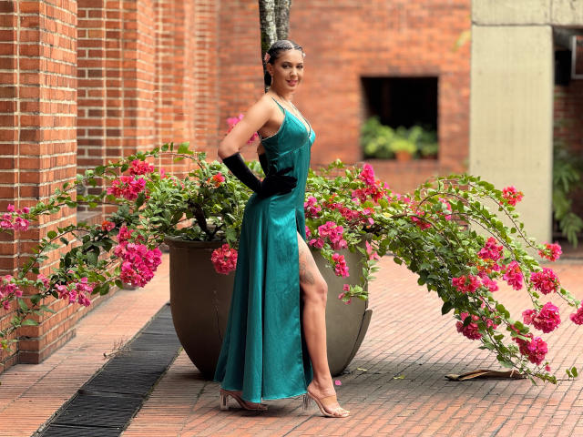 JoannaCool in a green, shiny, sleeveless dress with a high slit, black gloves, and high heels, standing next to a potted plant with pink flowers.